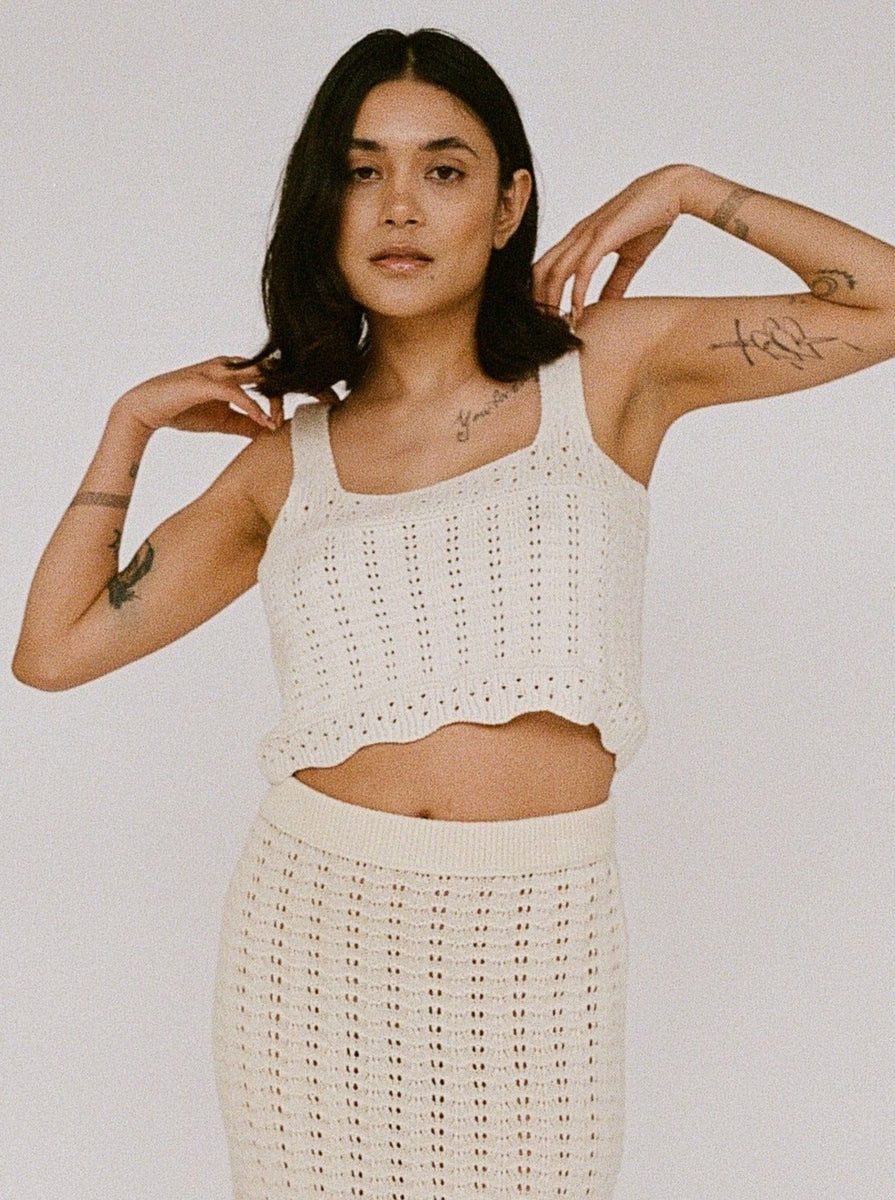 Sentence with replacement: Woman in a white Lyric Crochet Crop Tank - Ecru outfit, handmade in Peru, posing with hands raised slightly by her head.