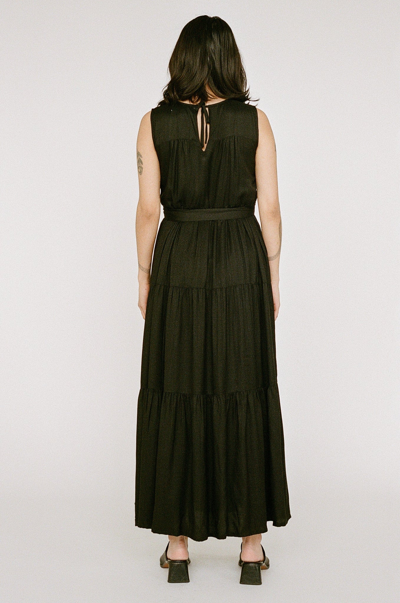 A person stands with their back to the camera, dressed in a Tiered Maxi Dress - Black made from Bemberg Moss Crepe and black heels, against a neutral backdrop.