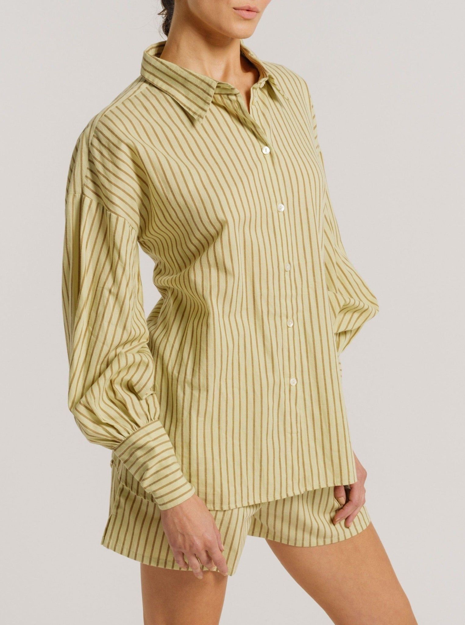 The model is wearing a Museo Button Up - Feather Grass Stripe shirt and shorts made from organic cotton.
