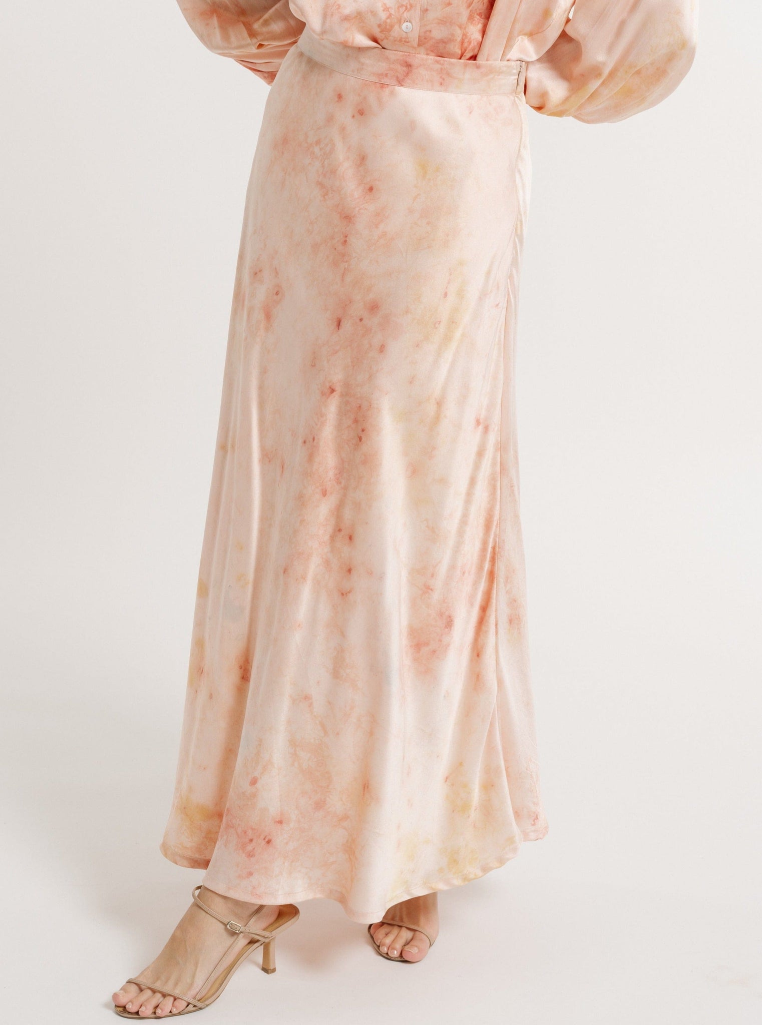 A person standing in a Peach-colored, Silk Midi Slip Skirt - Botanical Ice Dye with gold strappy high heels.