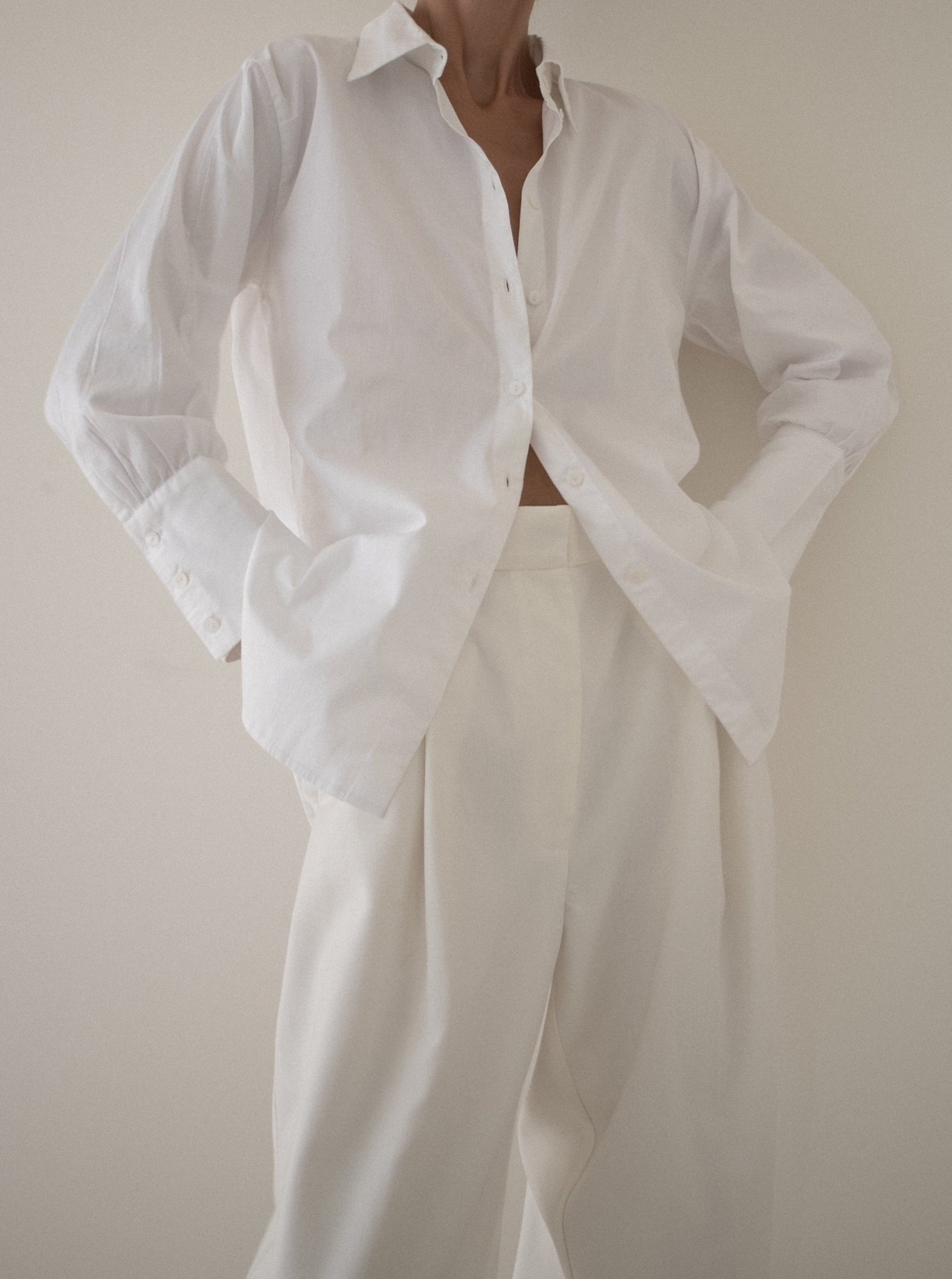 A woman wearing white pants and a white shirt, featuring mother of pearl buttons and made with organic cotton poplin for a slimmer fit, is wearing the Museo Button Up - White.