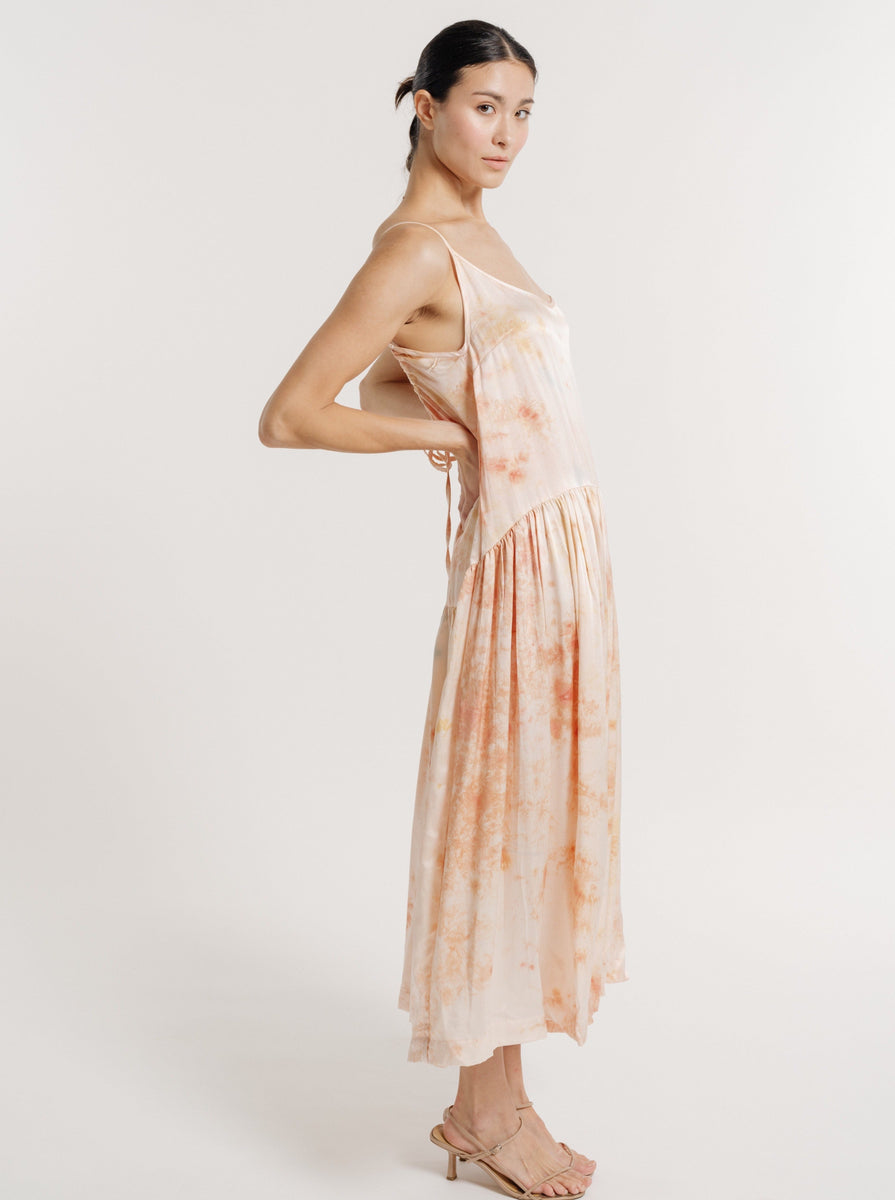 A woman standing sideways wearing a sleeveless Silk Midi Slip Dress - Botanical Ice Dye and sandals, looking over her shoulder at the camera.