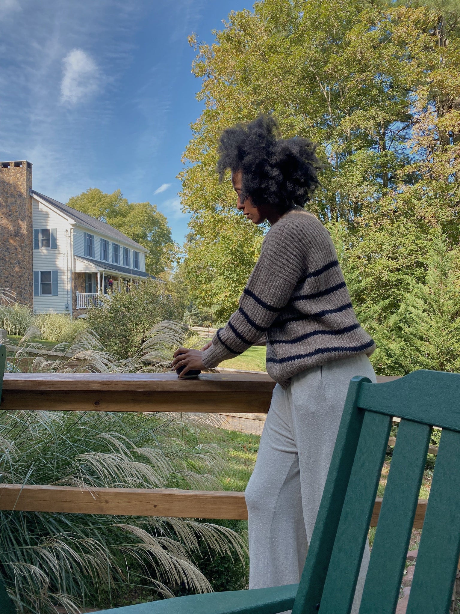 A woman sitting on a cozy green chair on a deck overlooking a house, dressed in a warm Field Sweater - Brown Stripe made of alpaca fiber.