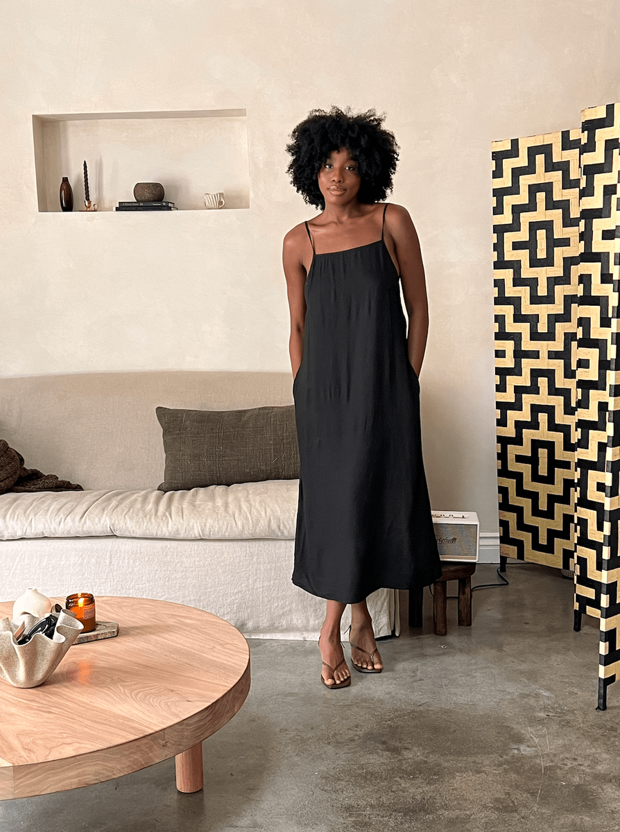 The sustainably made Kate Dress - Black, crafted from exquisite Bemberg Cupro Crepe, showcases a woman in a black dress standing in a living room.