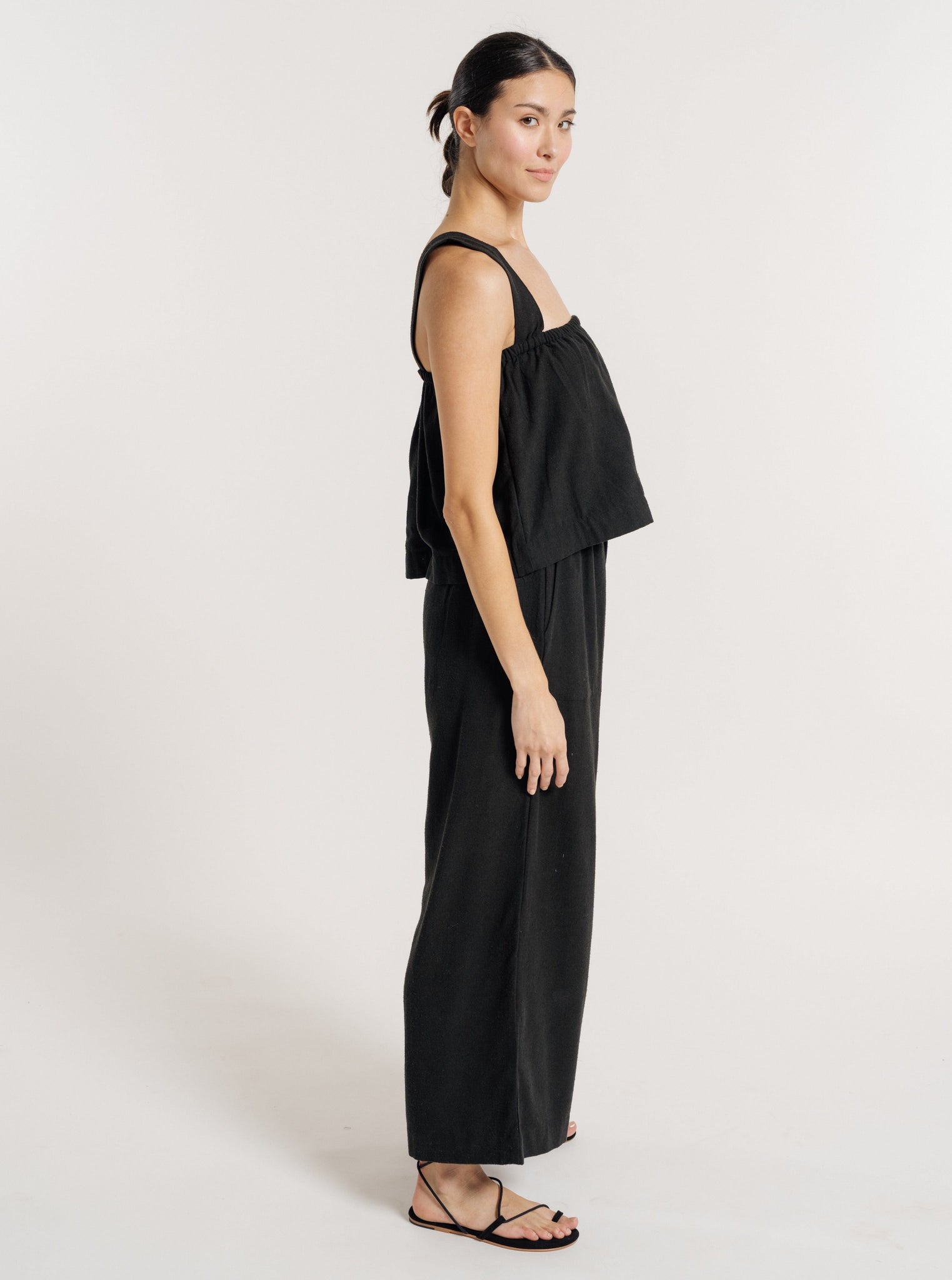 A woman wearing a Cross Back Tank - Black Silk Noil - Pre-order and sandals made of Silk Noil, perfect for resort pre-orders.
