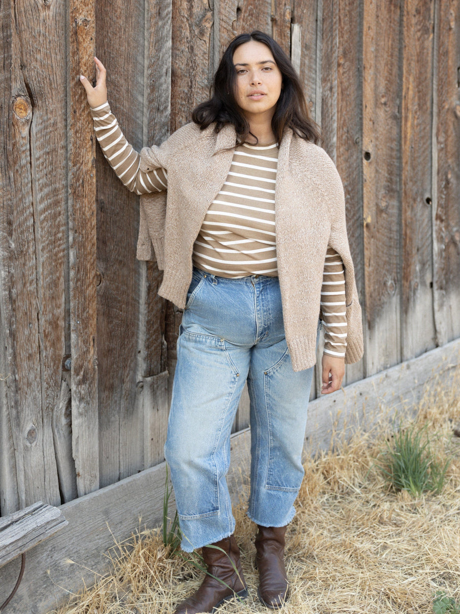 A woman wearing a Sample 144 - Mock Neck Long Sleeve Tee - Tannin Stripe and jeans leans against a wooden wall.