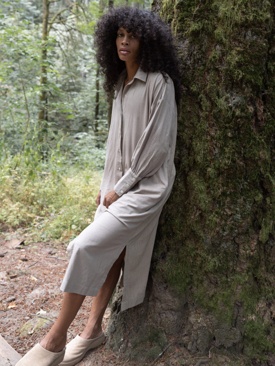 A black woman is leaning against a tree in a Dillon Dress - Mushroom - XXL - Sample.