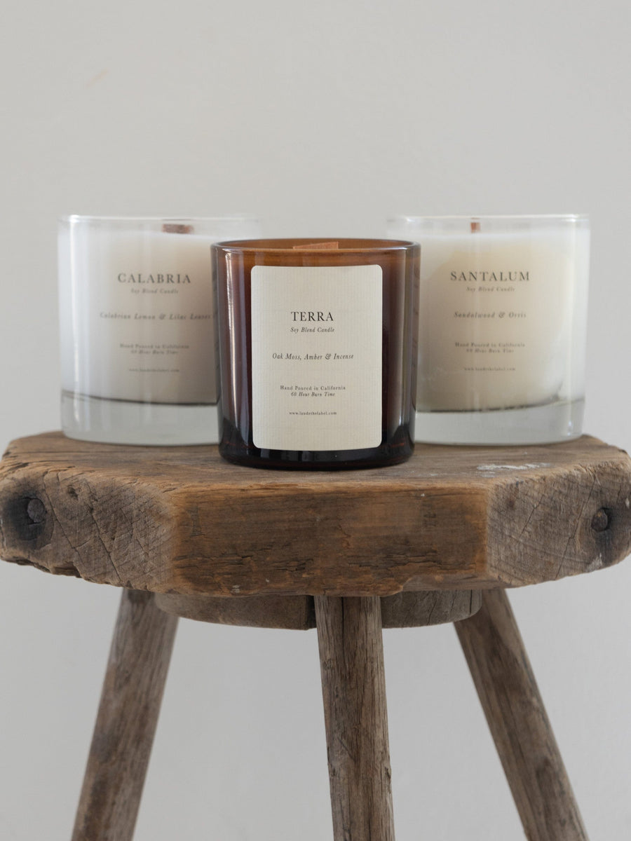Three Terra Glass Candles with wood wicks sitting on a wooden stool.
