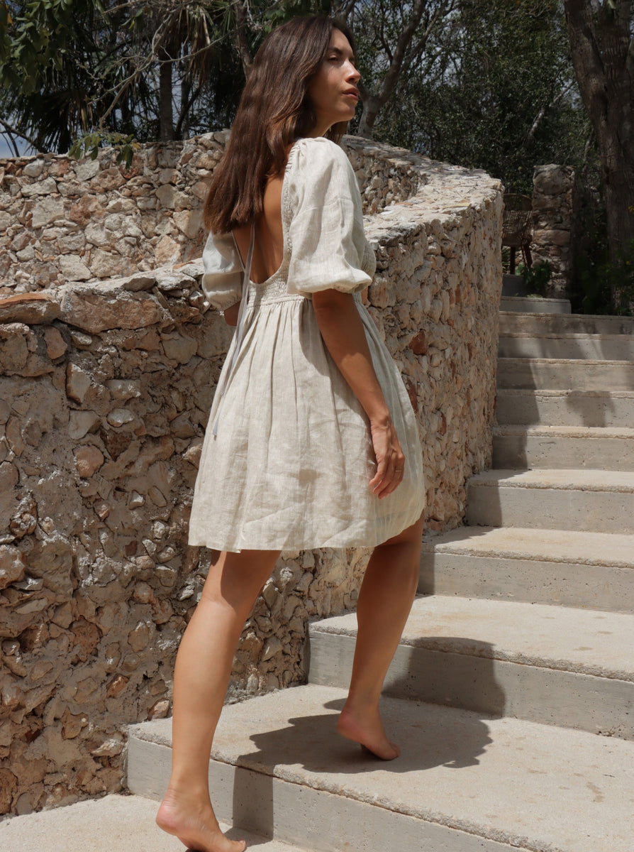 A woman in a beige Carmen Mini Dress - Natural - Sample gracefully descends a stone staircase.