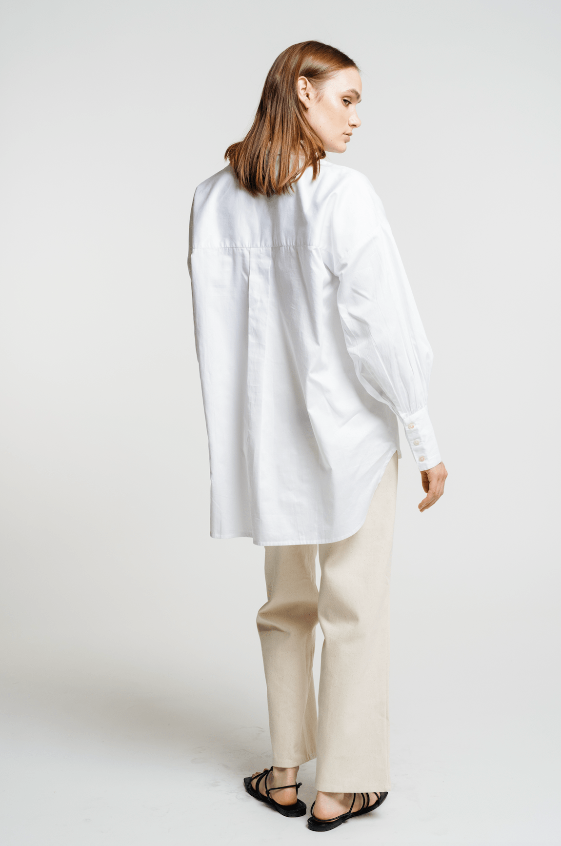 The back view of a woman wearing an Oversized Button Up - White, featuring an organic cotton poplin white shirt and beige pants.