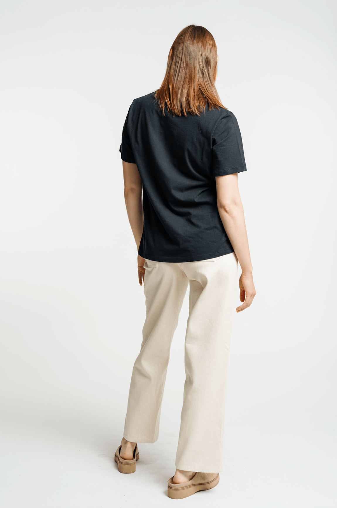 The woman is wearing an organic cotton black Crewneck T-Shirt and beige pants, seen from the back.