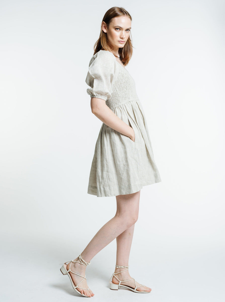 The model is wearing the Carmen Mini Dress - Natural - Sample in white with sandals, perfect for soaking up the Summer Sun. Made from Organic Linen.